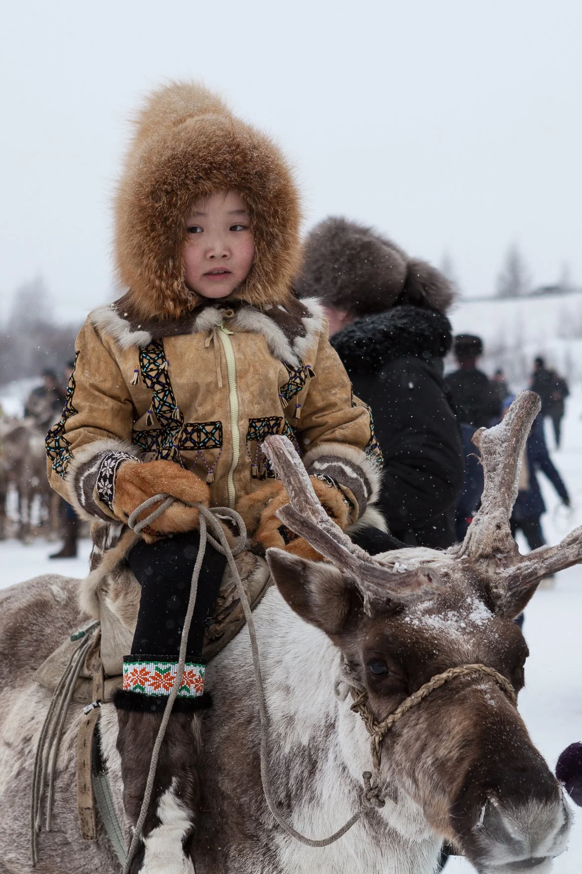 Enfant assis sur un renne, portant un manteau en laine et une capuche en fourrure, en hiver dans un paysage enneigé en Russie.