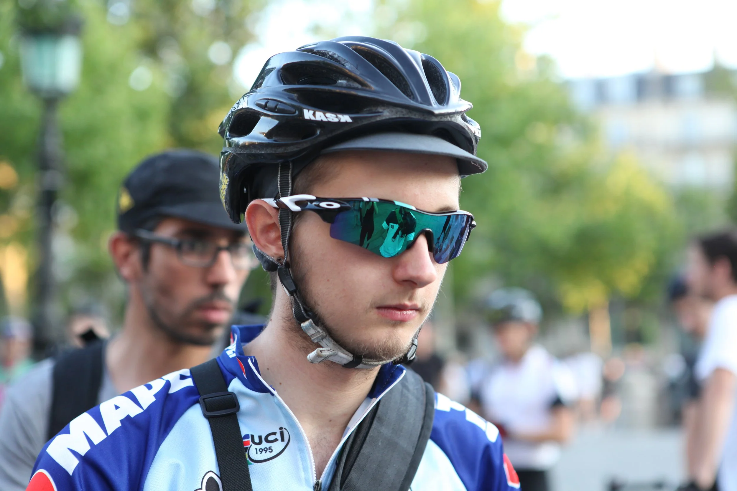 Un jeune homme porte un casque de vélo, des lunettes de soleil sportives et une veste de cyclisme avec des logos, à l'extérieur avec un groupe de personnes en arrière-plan.