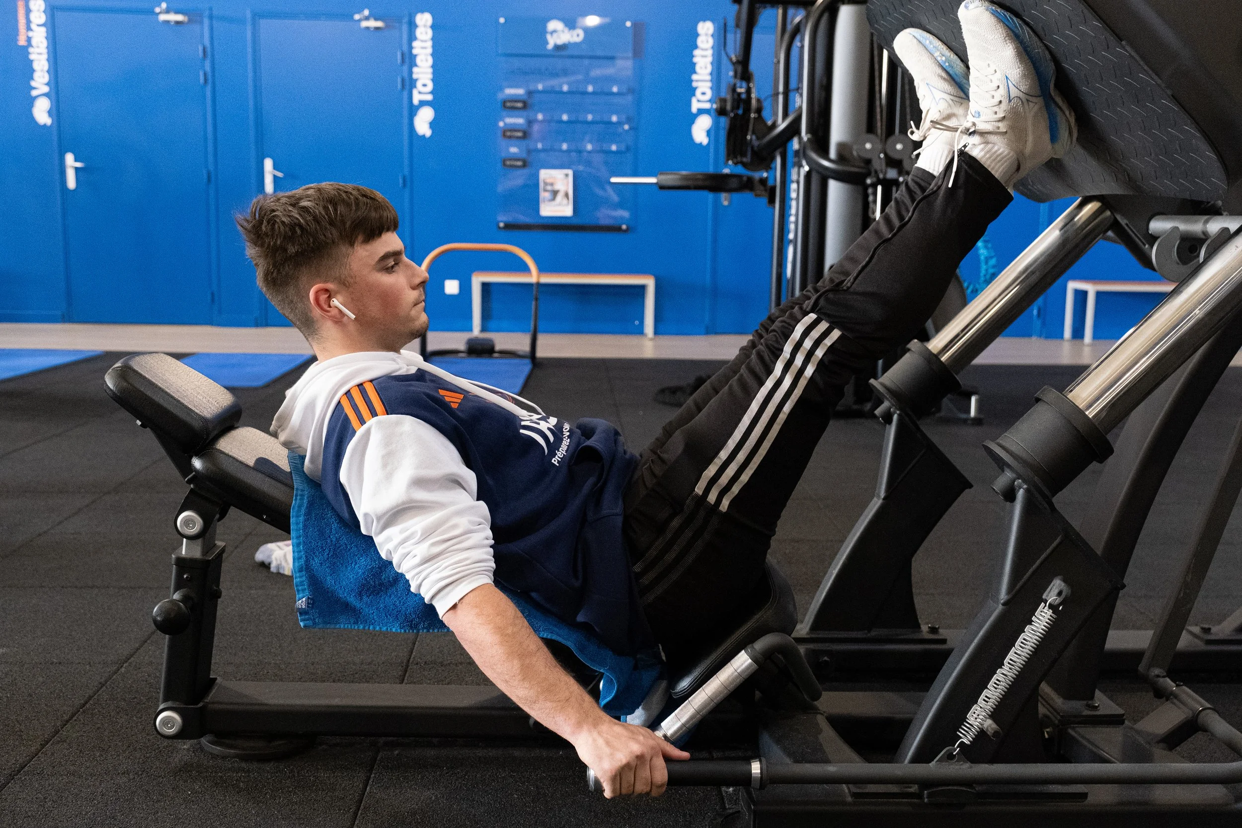 Un jeune homme s'entraîne à faire du leg press dans une salle de sport, portant des écouteurs et un sweat à capuche.