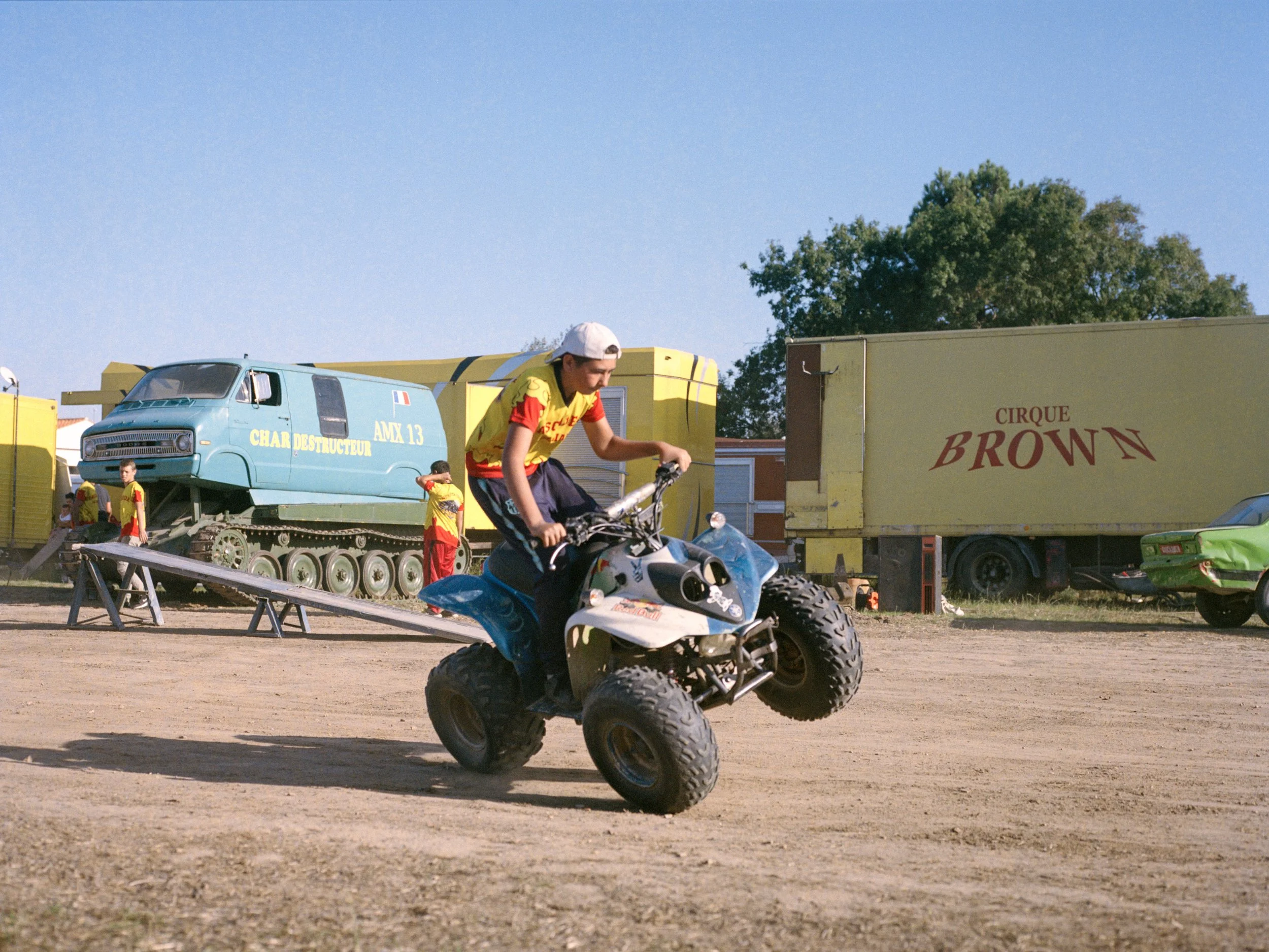 Un jeune homme fait une figure sur un quad lors d'un show de Monster Truck, avec des camions du cirque 'Brown' en arrière-plan.