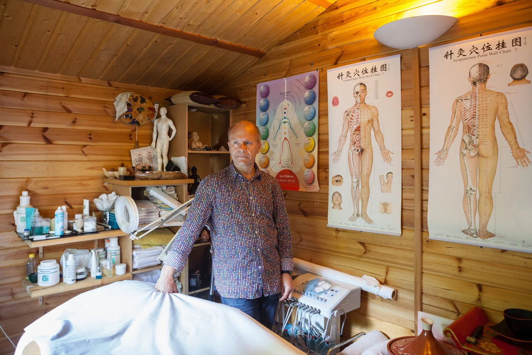 Un homme dans une pièce avec des murs en bois, entouré de posters sur la médecine traditionnelle chinoise et des étagères avec des produits et des objets de décoration.