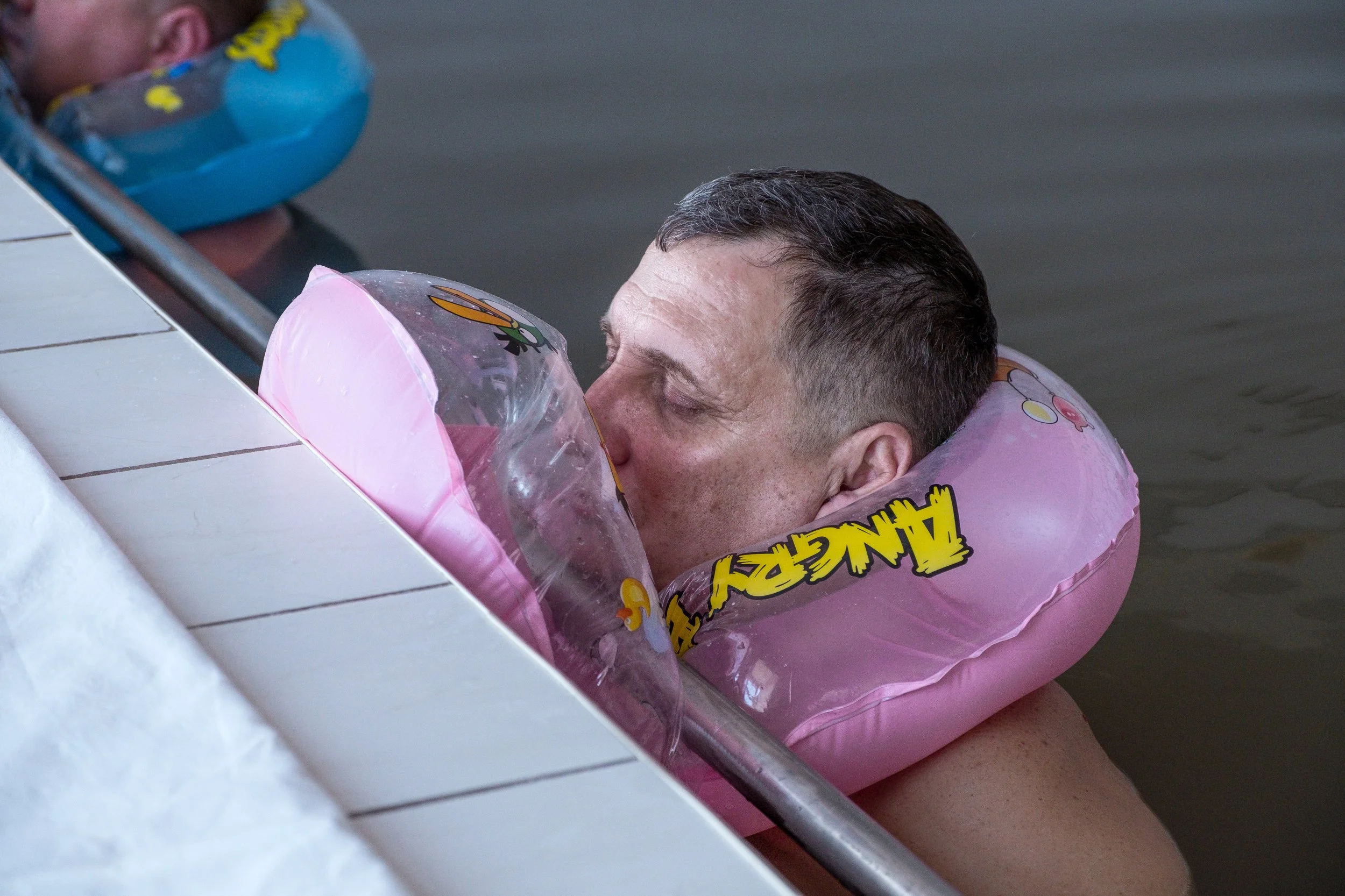 Un homme nageant dans une piscine, portant une bouée de flotteur rose avec des dessins et le mot 'ANGRY' écrit dessus.