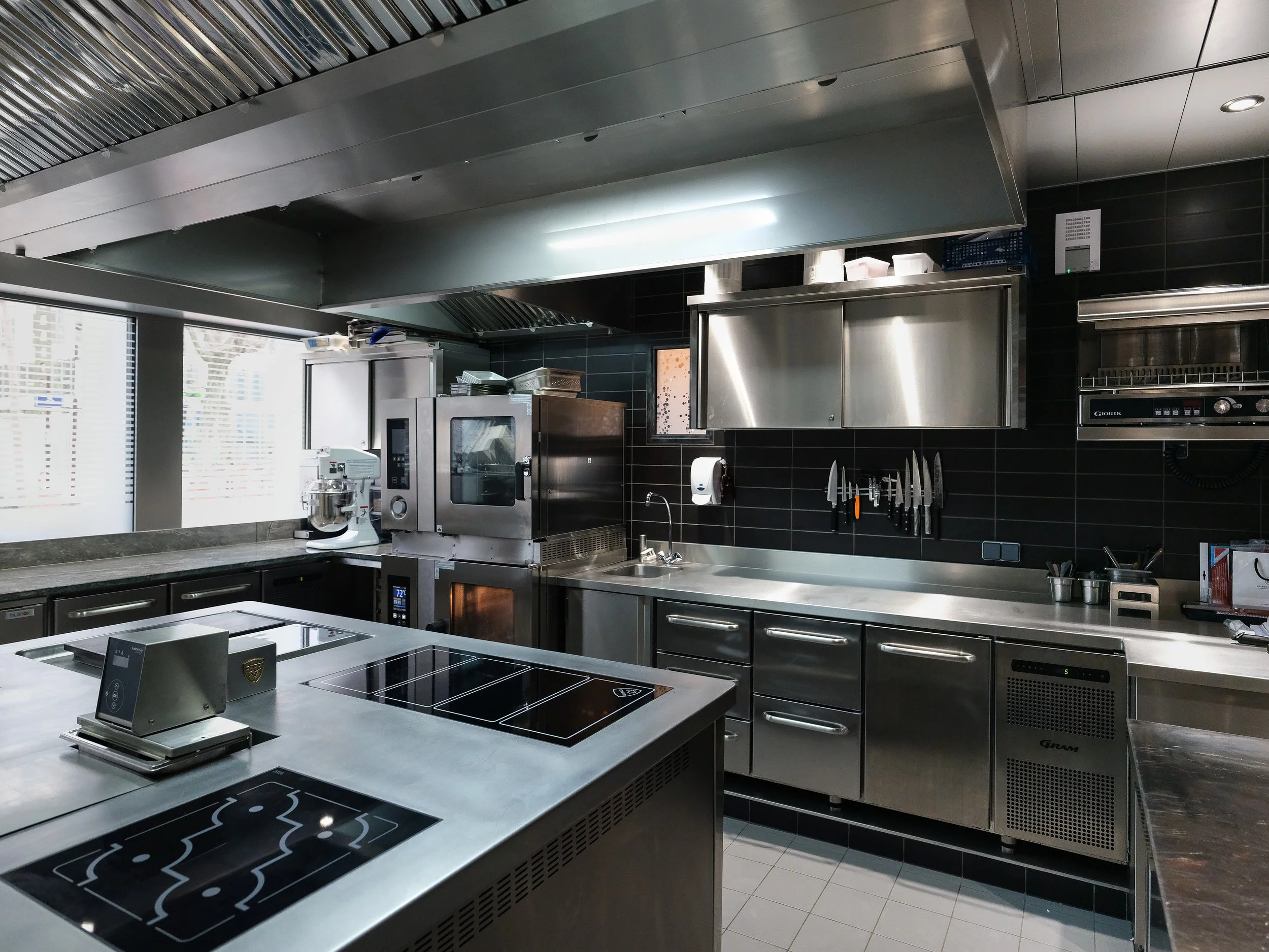 Cuisine professionnelle en acier inoxydable avec four, plaques de cuisson, mitigeur, couteaux accrochés au mur, et divers équipements de cuisine.