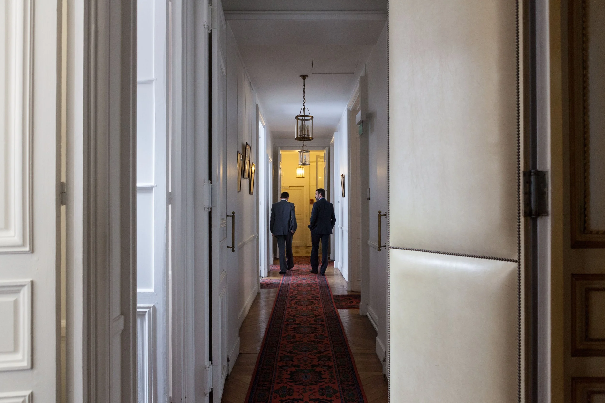 Deux hommes en costume marchant dans un couloir de l'Elysée, éclairage tamisé avec lampes suspendues, murs décorés avec quelques tableaux.