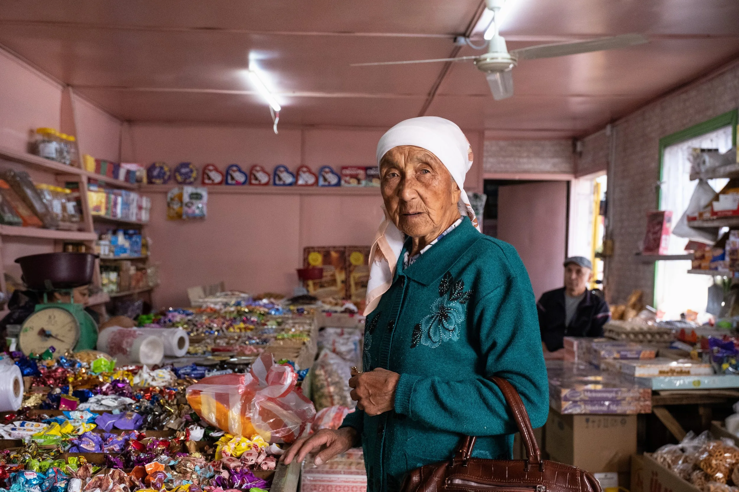 Une femme âgée portant une coiffe blanche et un manteau vert, qui fait du shopping dans une boutique remplie de bonbons et de produits emballés. En arrière-plan, un jeune homme au fond du magasin au Kazakhstan.