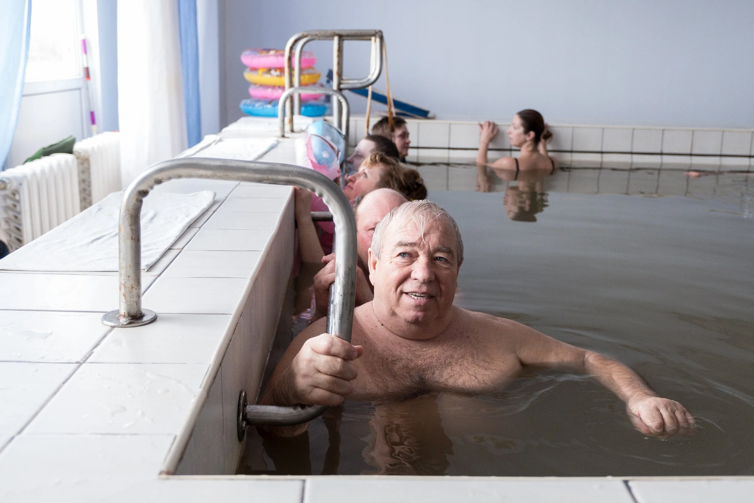 Groupe de personnes âgées et jeunes dans une piscine intérieure, certaines accrochées au rebord, d'autres nageant ou debout dans l'eau, arrière-plan avec des tubes de natation.