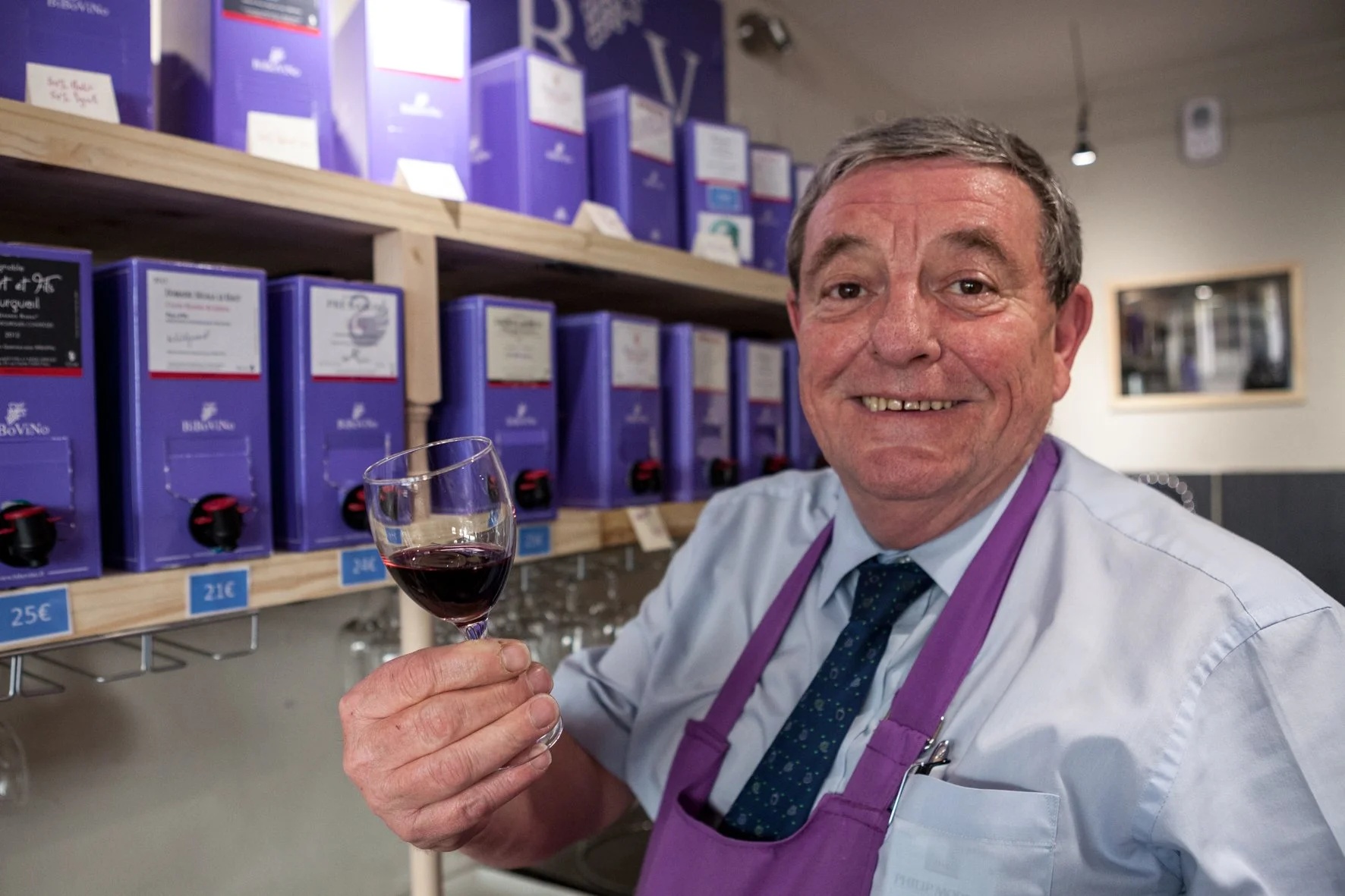 Un homme souriant tient un verre de vin rouge dans une cave ou un magasin de vins, avec des étagères remplies de bouteilles de vin en arrière-plan.