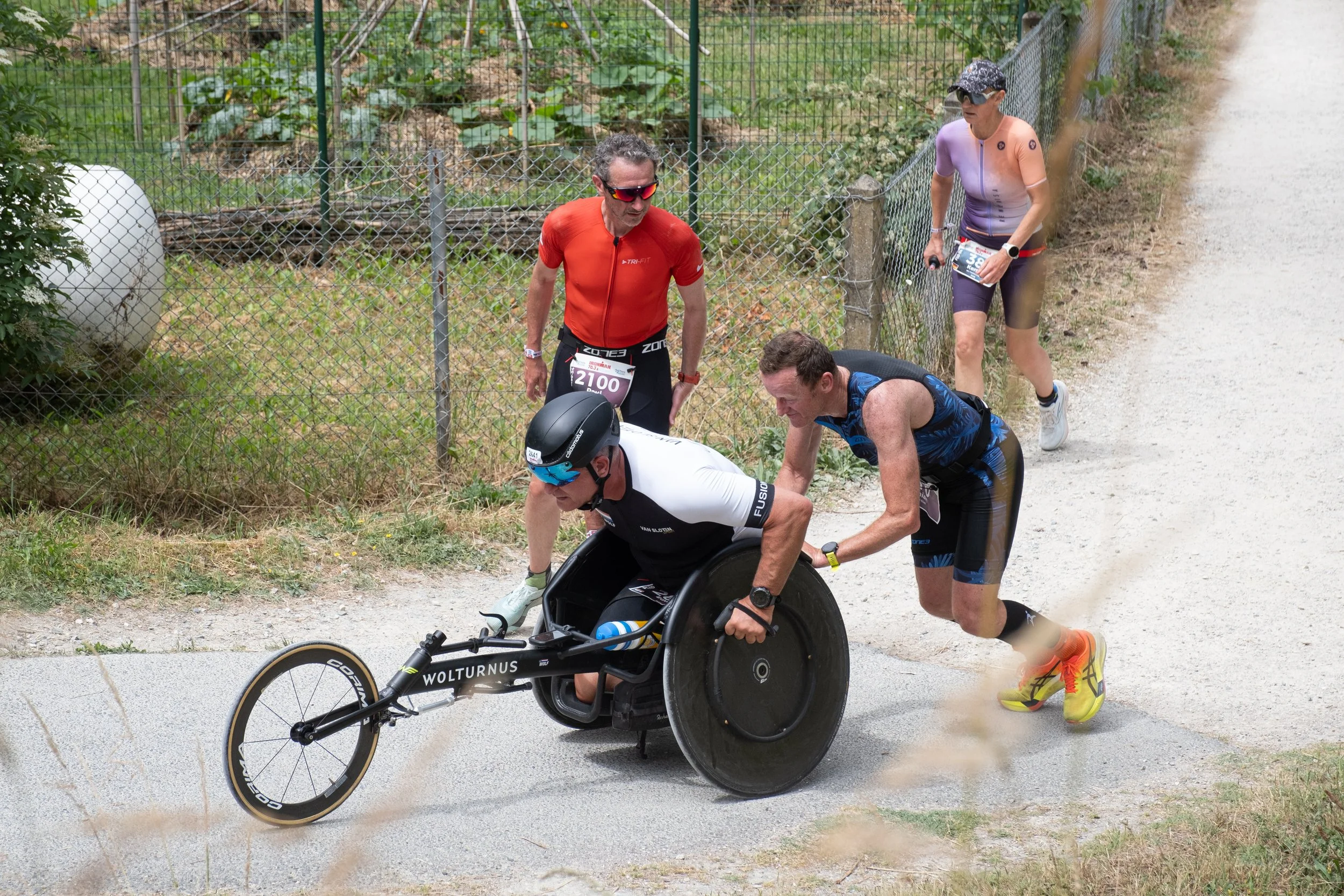 Un athlète en triathlon en fauteuil roulant reçoit une assistance sur le parcours, entouré de trois autres participants.