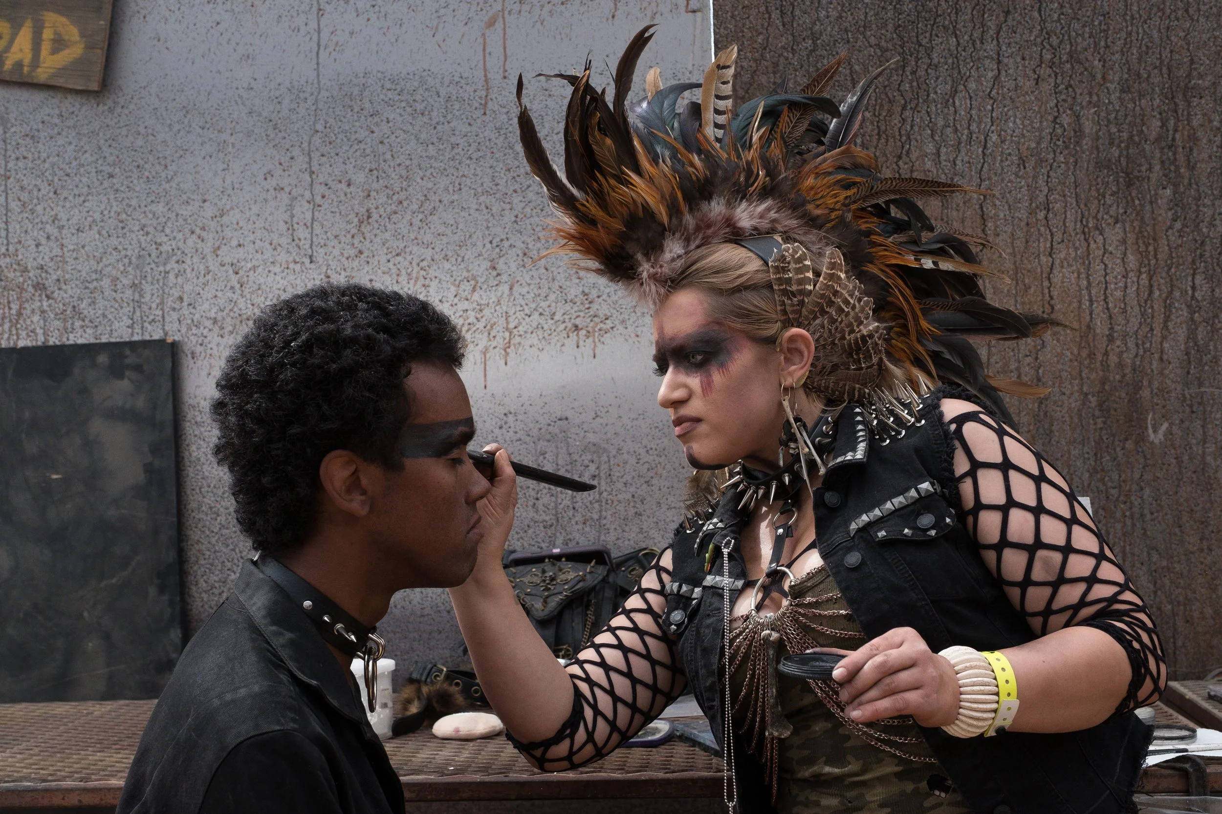 Une femme en déguisement punk avec un grand couvre-chef en plumes et des vêtements cloutés applique du maquillage à un jeune homme.