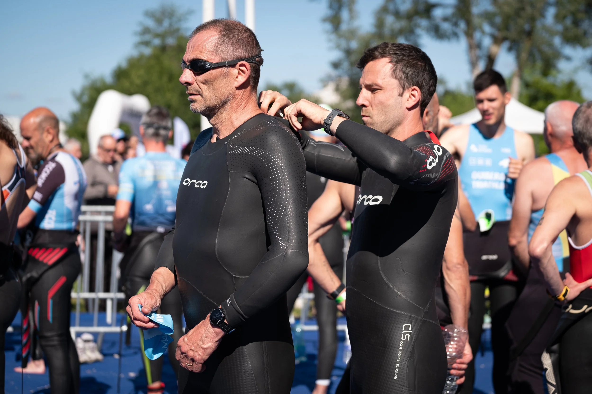 Deux hommes en tenue de triathlon noir se préparent avant la course, l'un ajustant la peau de l'autre, entourés d'autres triathlètes en arrière-plan, lors d'une compétition en plein air sous un ciel ensoleillé.