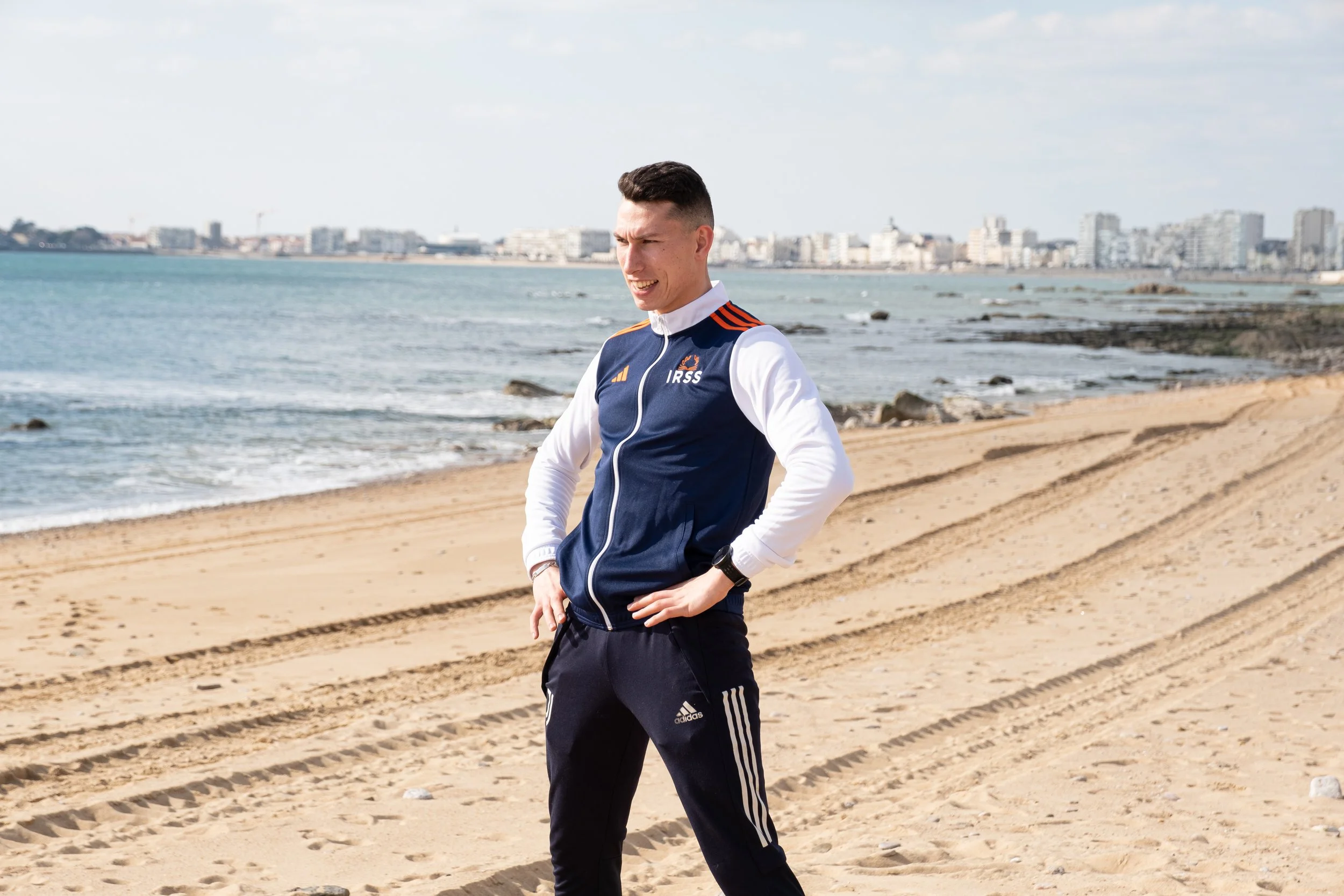 Un jeune homme en tenue de sport dans une plage de sable, regardant au loin avec un arrière-plan d'immeubles près de la mer.