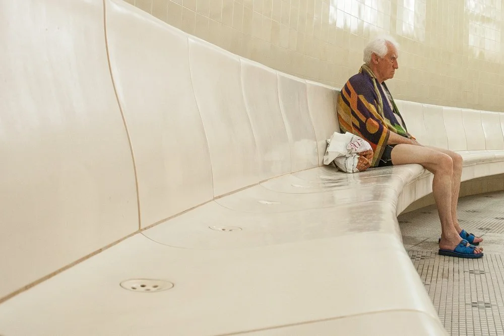 Un homme âgé assis seul sur un banc en céramique aux bains Szechenyi de Budapest, portant une serviette de bain colorée sur ses épaules, un maillot de bain et des sandales bleues.