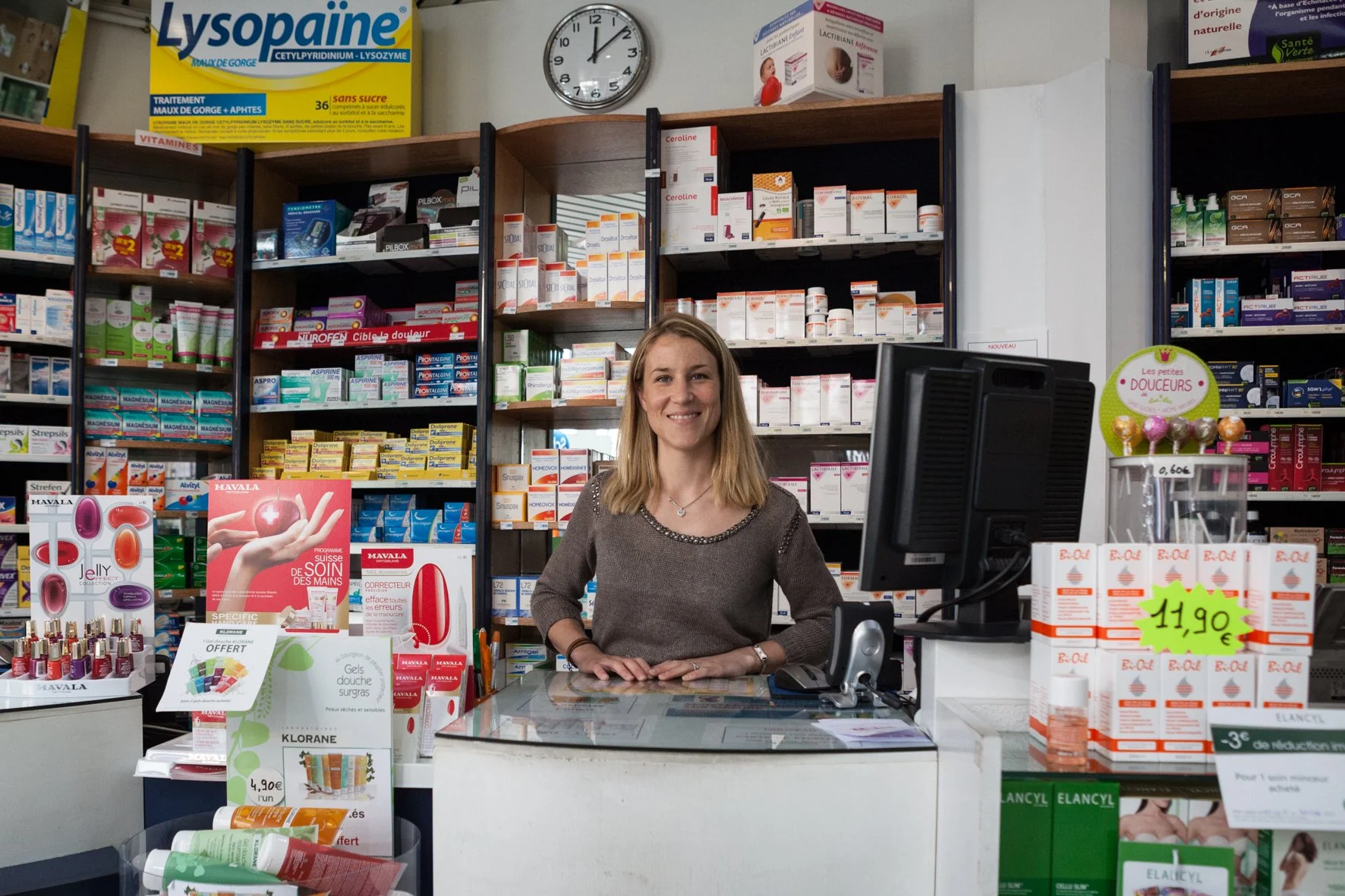 Une femme avec un sourire derrière un comptoir dans une pharmacie ou une parapharmacie, avec des étagères remplies de produits de santé et de beauté en arrière-plan.