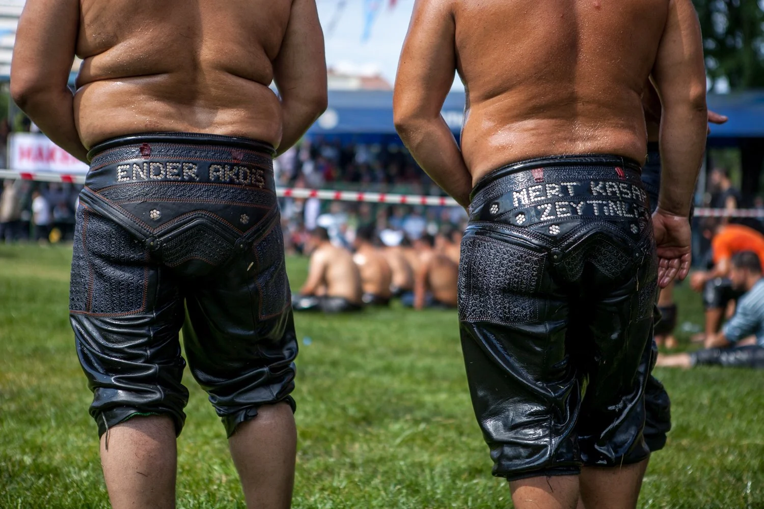 Deux hommes debout, portants des pantalons en cuir noir, lors d'un événement en plein air. Leurs noms sont brodés au dos de leurs pantalons.