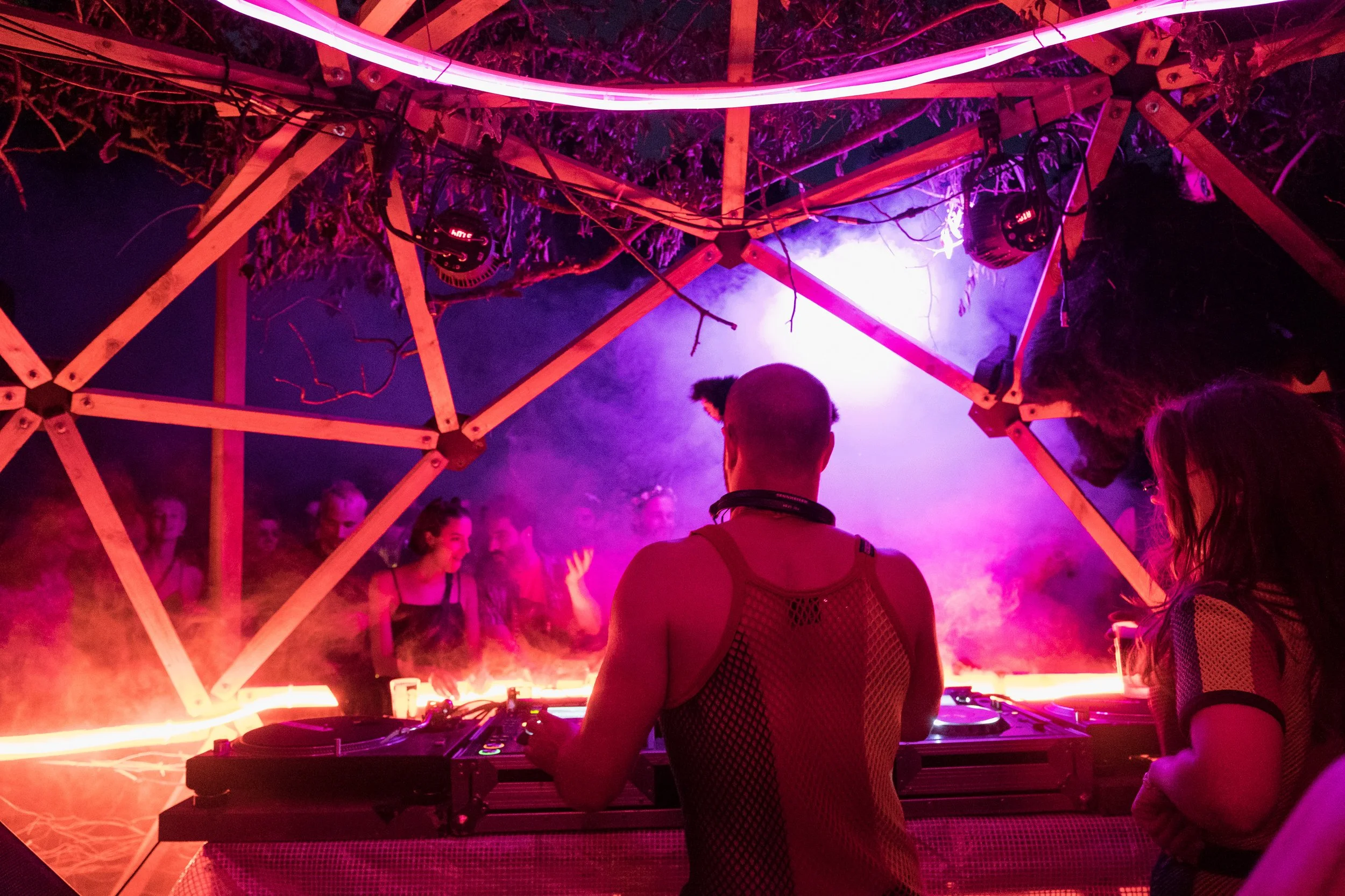 DJ jouant dans le festival en plein air de Lapiñata avec décor en bois et éclairage lumineux violet et rose, entouré de plusieurs personnes dansant et socialisant.