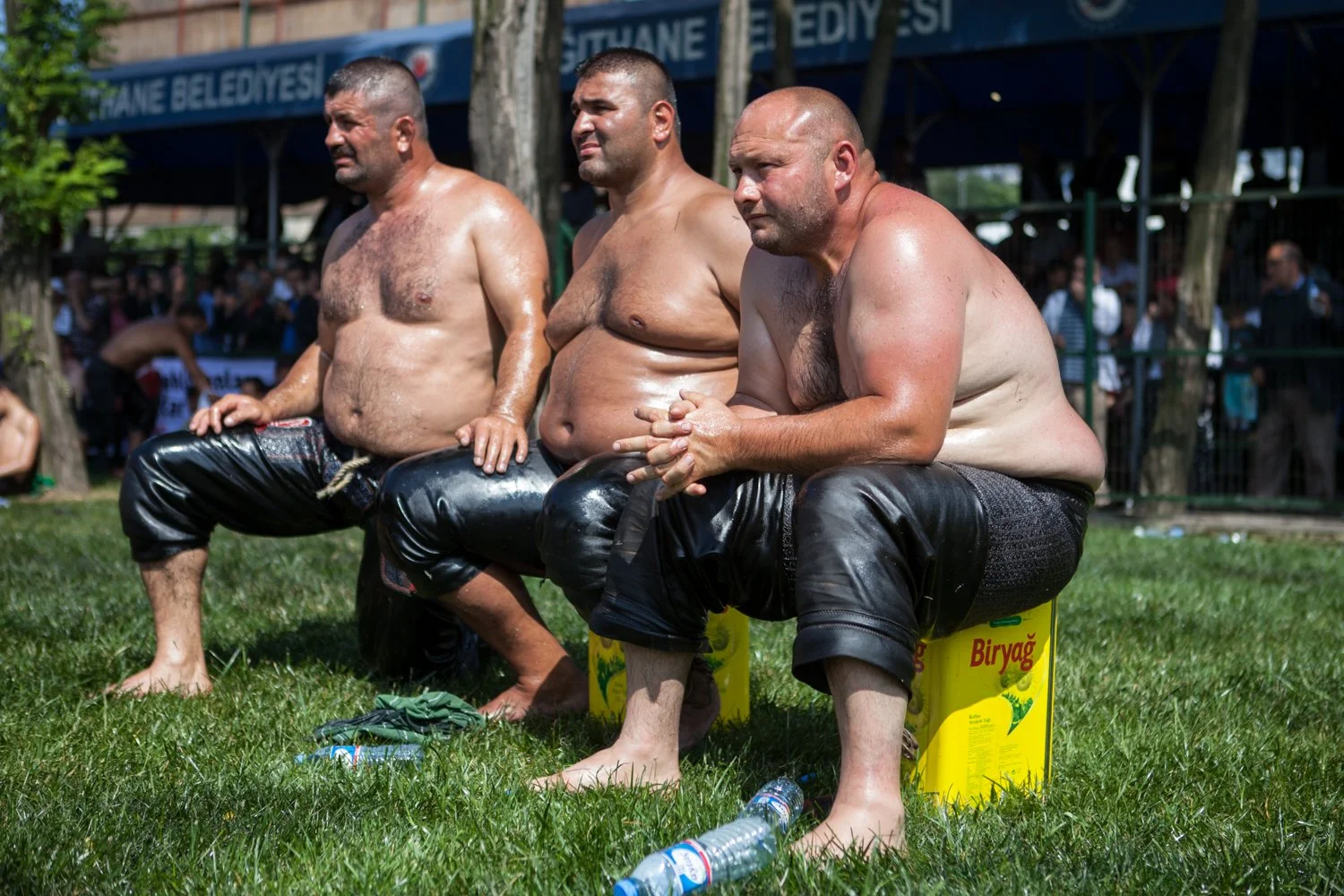 Trois hommes nus, assis sur des boîtes de Biryag, lors d'un événement public en plein air, avec un public en arrière-plan.
