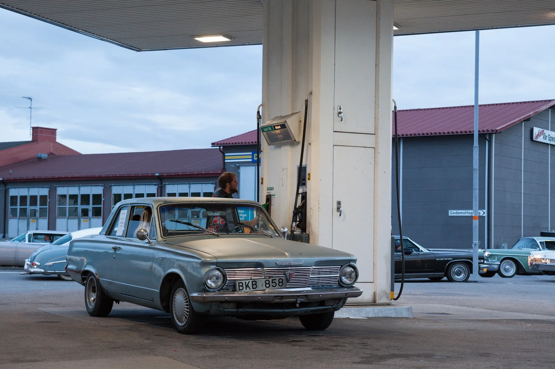Une voiture vintage gris clair fait le plein à une station-service. Un homme avec une barbe et une veste sombre se trouve à la pompe, avec plusieurs autres voitures classiques visibles en arrière-plan.