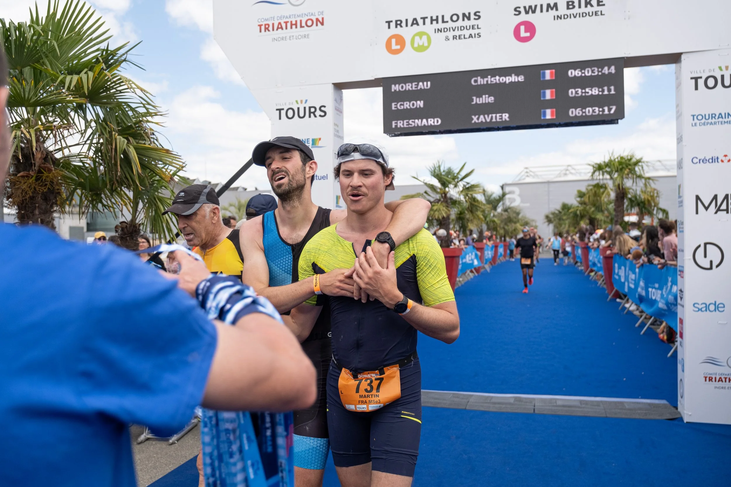Des coureurs terminent une course de triathlon, avec un panneau d'arrivée derrière eux indiquant les temps de parcours et les noms. La scène se déroule en plein air avec un ciel partiellement nuageux et des palmiers.