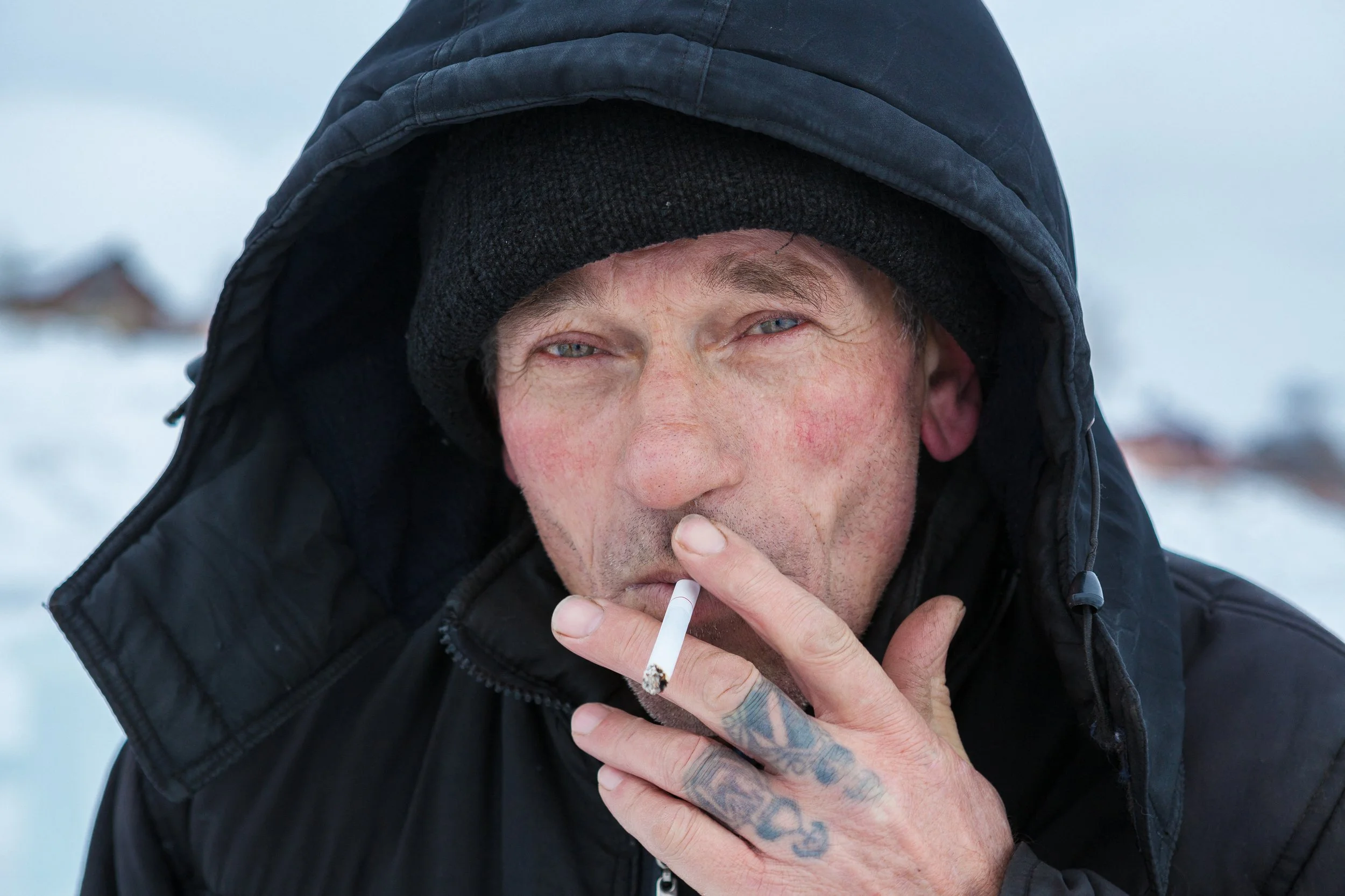 Un homme portant une capuche et un manteau noir, tenant un cigarette, dans un environnement enneigé.