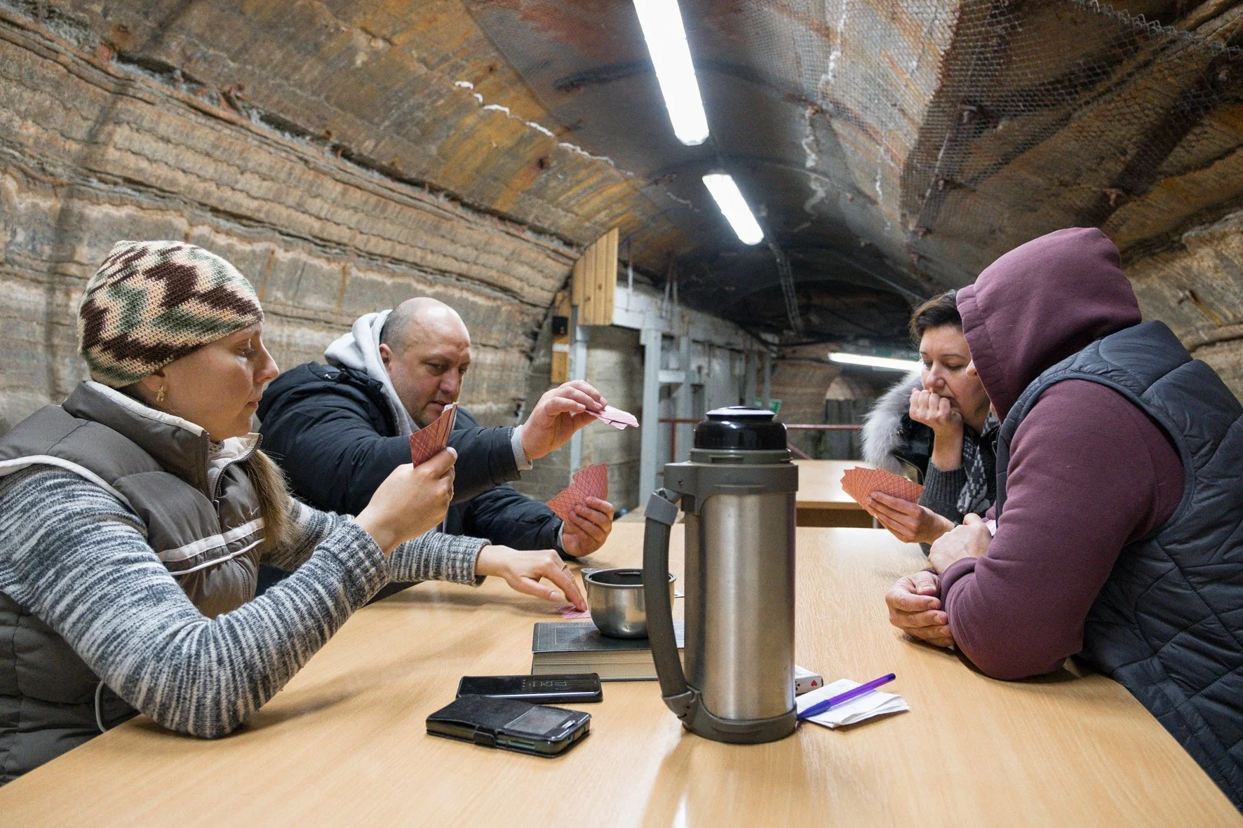 Quatre personnes jouent aux cartes dans un tunnel souterrain en utilisant une table en bois et un thermos, ambiance détendue.