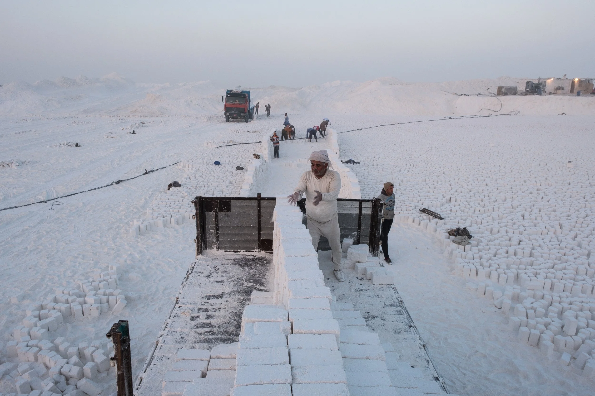 Homme maniant des blocs de calcaire dans un paysage tout blanc, avec plusieurs autres personnes travaillant à distance dans le fond.