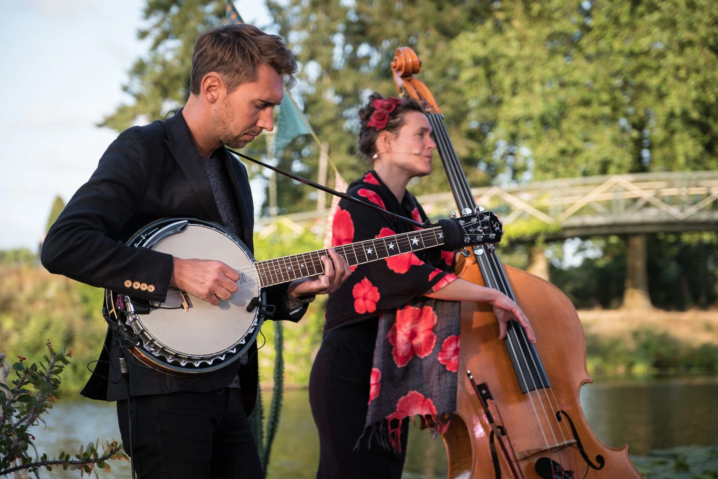 Deux musiciens au festival de la rive aux Barges jouent de la musique en plein air, l'un avec une guitare banjo et l'autre avec une contrebasse, dans un environnement naturel avec des arbres et un pont en arrière-plan.