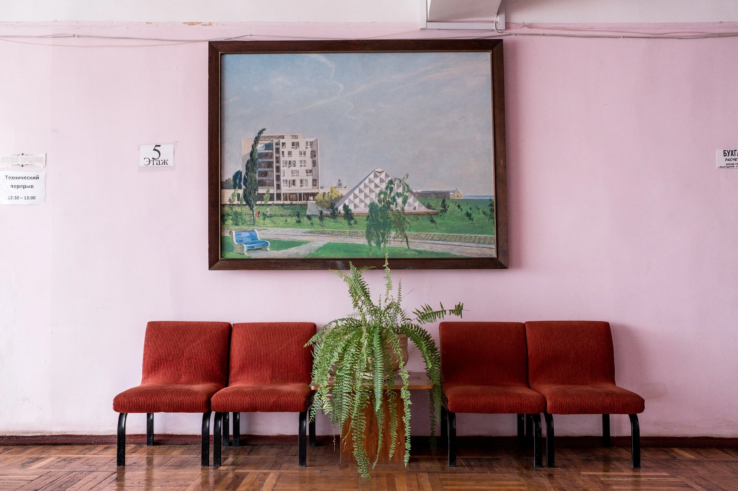Salle d'attente avec quatre chaises rouges, un pot de plante verte, une peinture représentant un bâtiment et un parc sous un ciel bleu à Kuyalnik, en Ukraine.