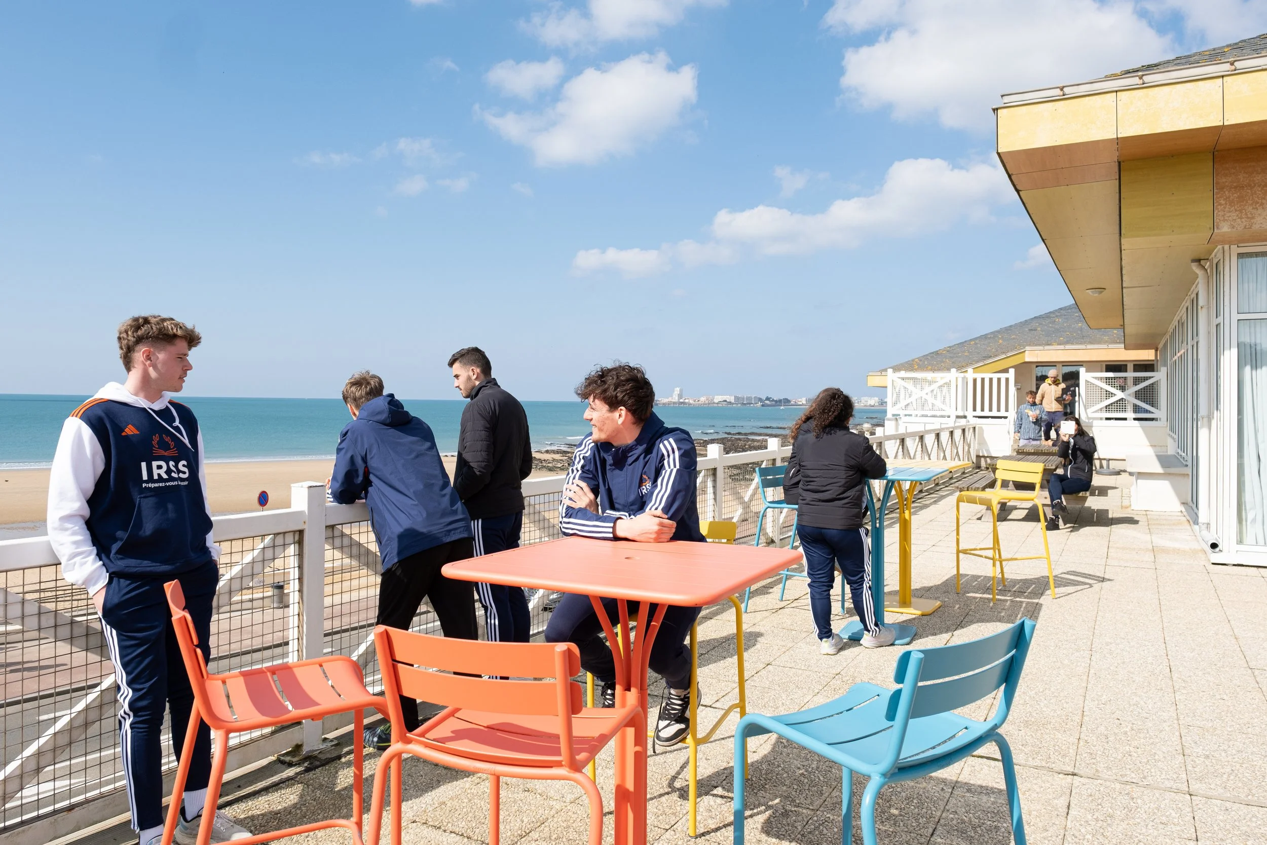 Groupe de personnes assises et discutant sur une terrasse face à la mer, avec des chaises colorées et un ciel ensoleillé.