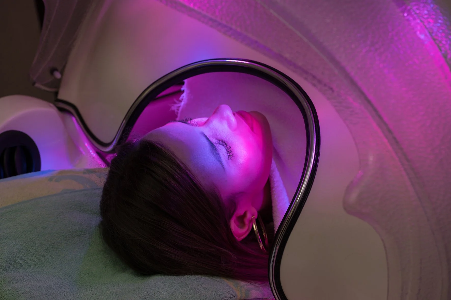 Une personne allongée sous une machine de luminothérapie, avec des lumières colorées roses et violet. La personne porte des boucles d'oreille et a les yeux fermés.