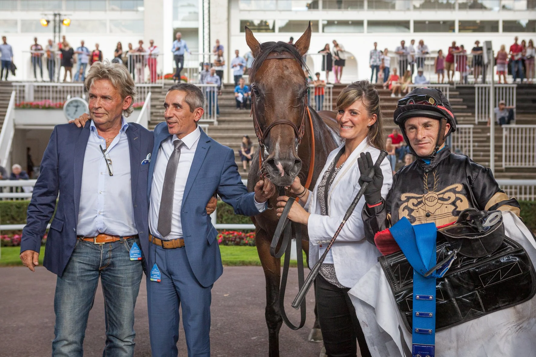 Groupe de personnes avec un cheval au centre, en compétition hippique, avec des spectateurs dans les gradins en arrière-plan.