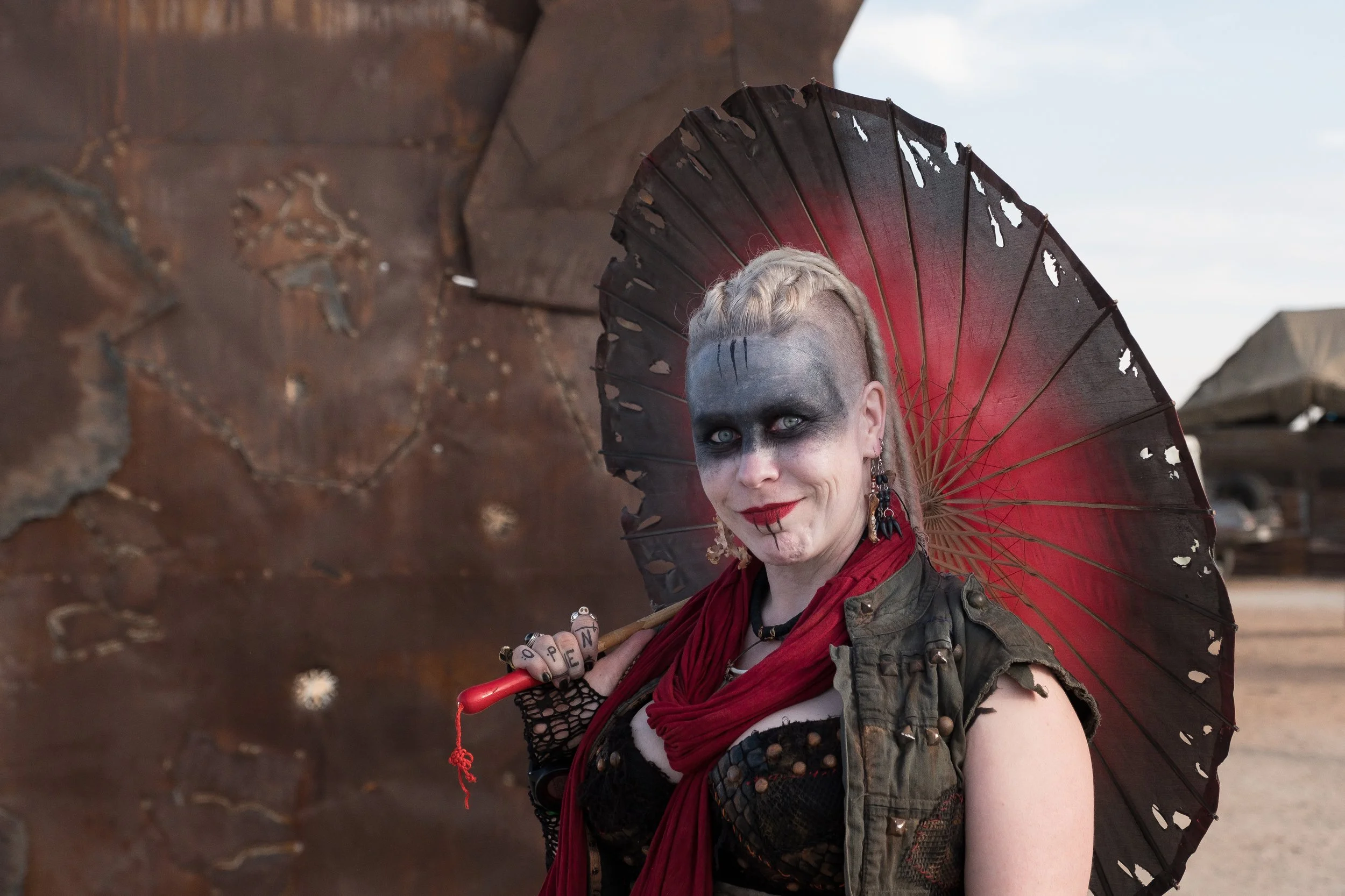 Une femme portant un maquillage sombre et un parapluie noirâtre avec des trous, vêtue d'une veste usée et d'un foulard rouge, se tenant dans un paysage désertique avec un ciel clair.