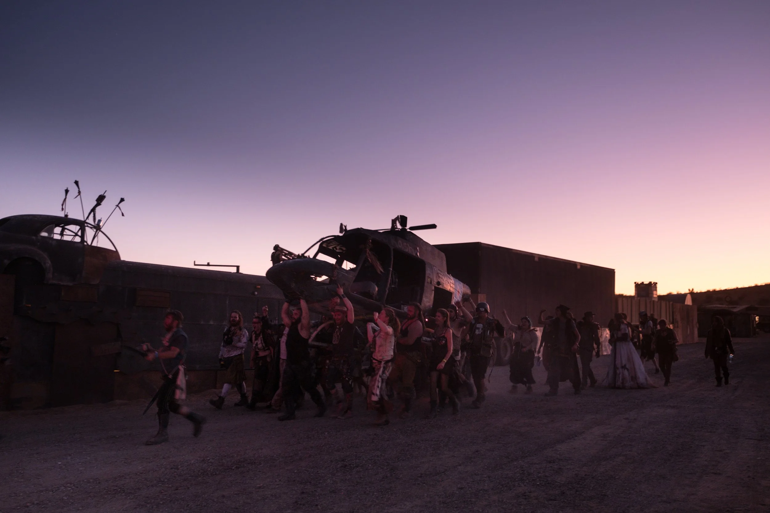 Groupe de personnes déguisées marchant dans un décor désertique et portant un hélicoptère sur leurs épaules à la tombée de la nuit à Wasteland.