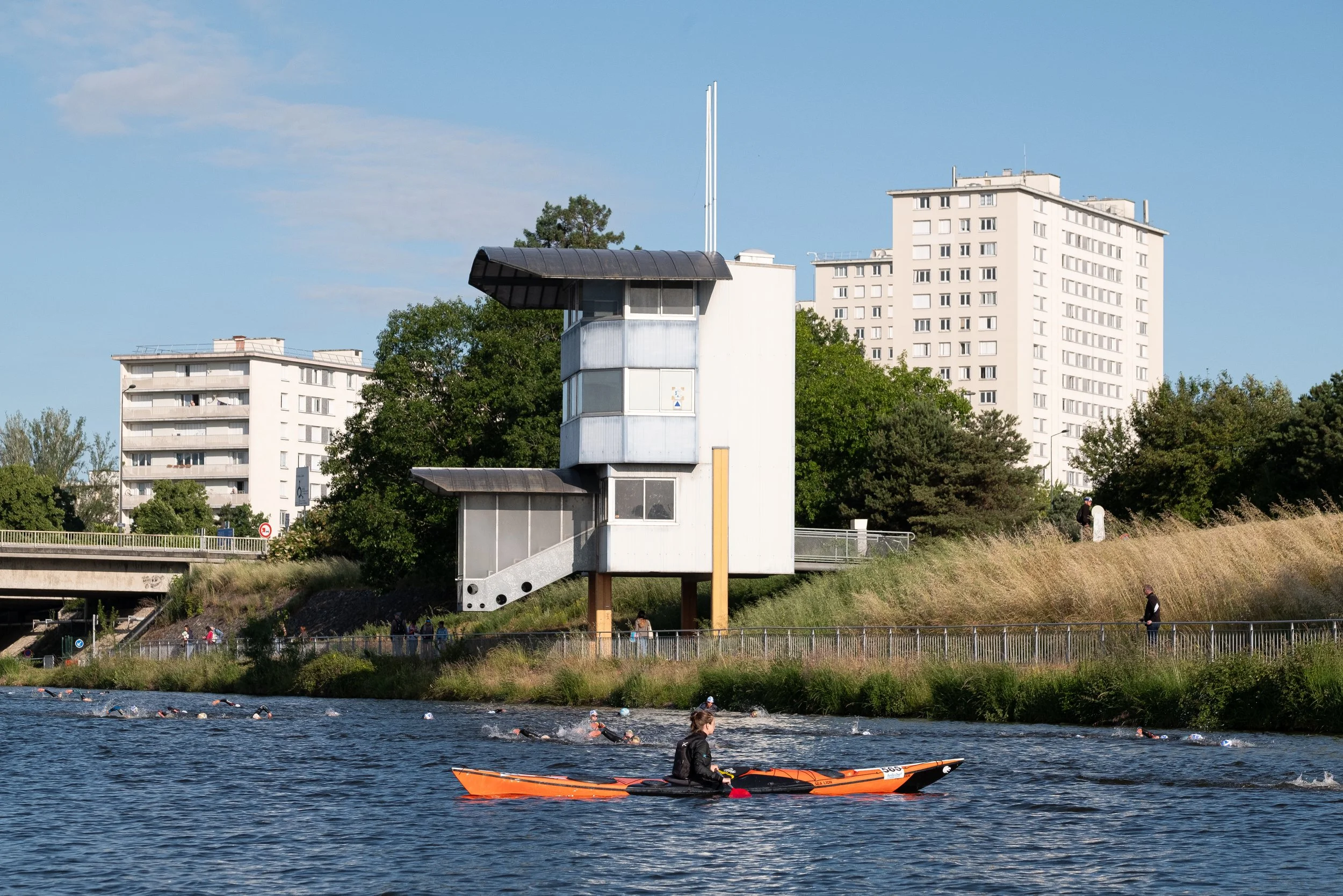 Un kayakiste qui navigue sur une rivière, avec un bâtiment moderne en arrière-plan et des immeubles résidentiels