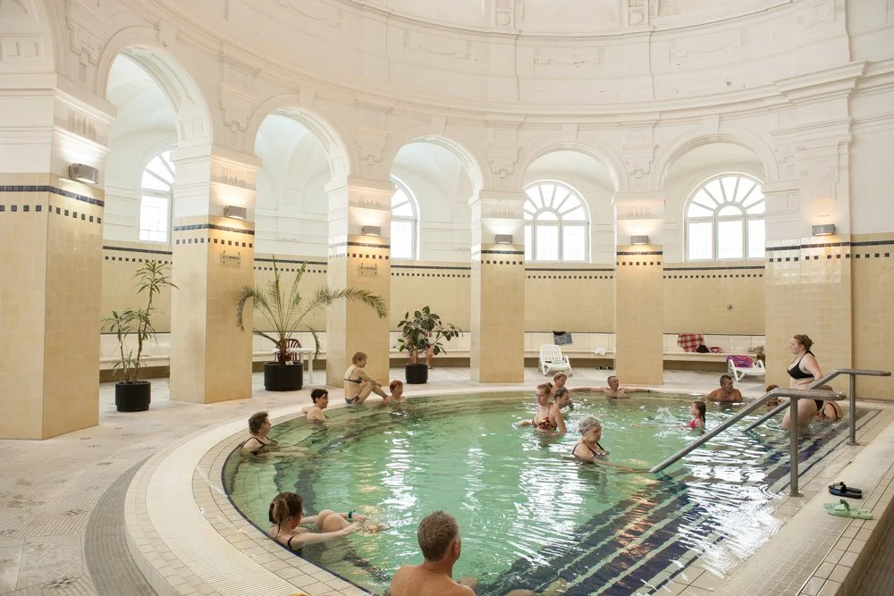 Piscine intérieure de Szechenyi à Budapest avec plusieurs personnes se relaxant, dans une salle lumineuse avec fenêtres arquées, plantes et mobilier de détente.