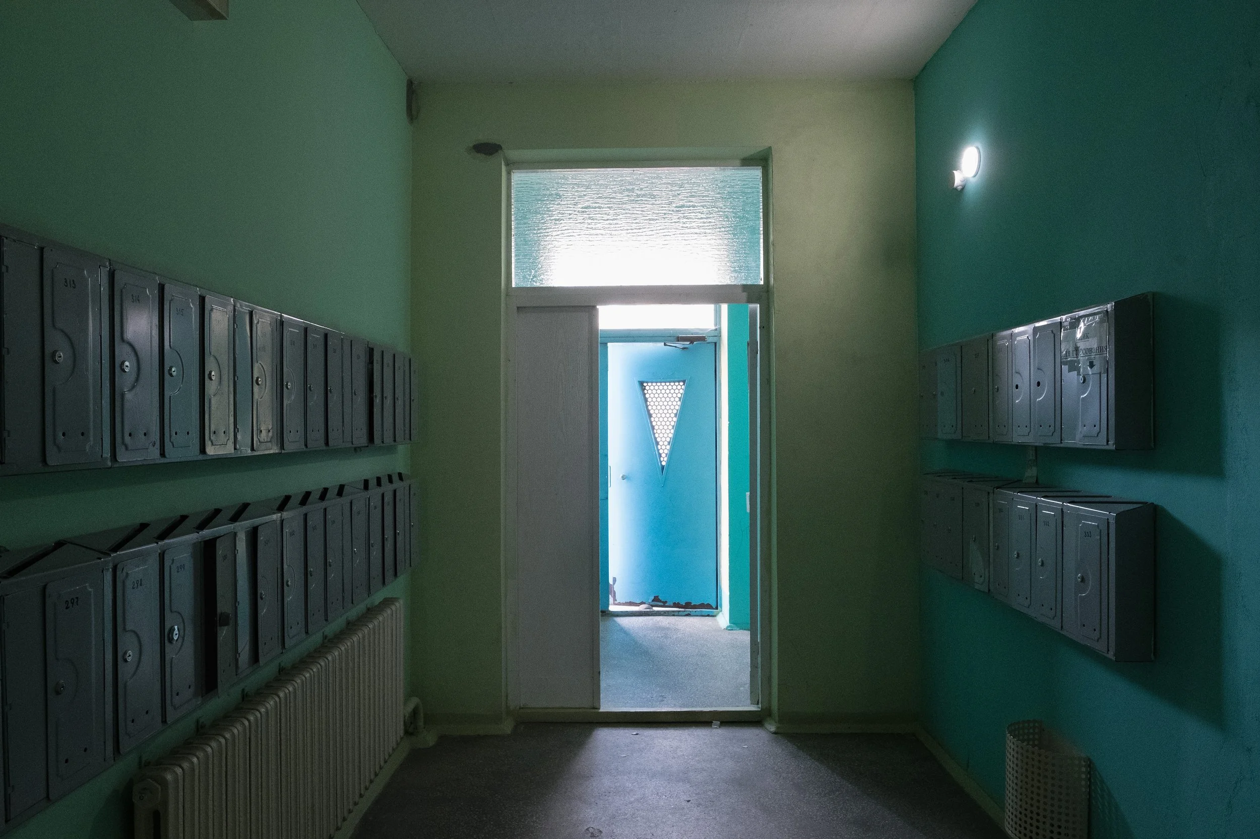 Couloir d'immeuble avec des boîtes aux lettres en métal gris sur les murs vert pâle, porte d'entrée bleue au fond avec une fenêtre en haut, et un radiateur blanc.