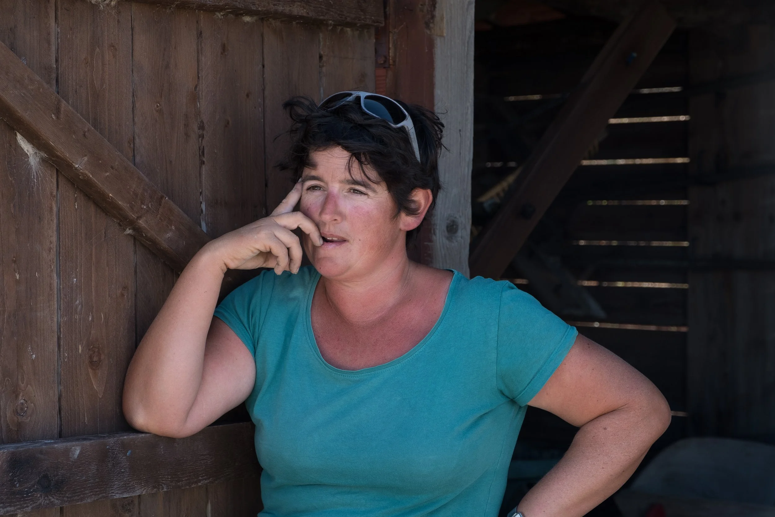 Une femme en t-shirt turquoise, portant des lunettes de soleil sur la tête, pose contre un mur en bois. Elle a l'air pensive, avec une main sur la joue.