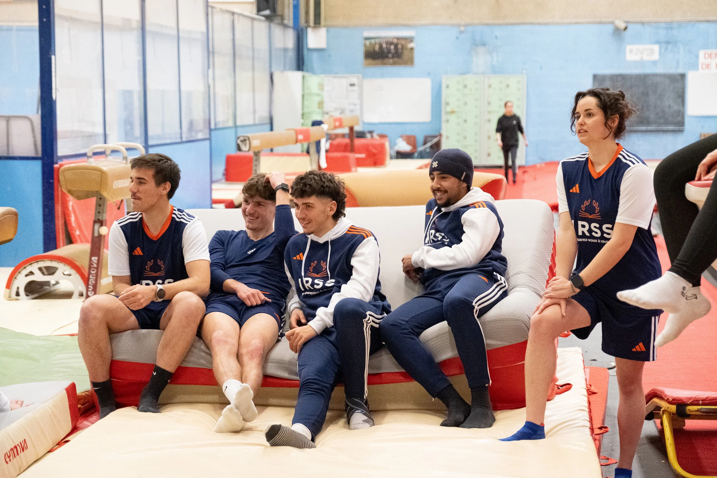 Groupe de six jeunes sportifs assis sur un canapé dans une salle d'entraînement de gymnastique, portant des vêtements de sport à l'effigie de leur club.