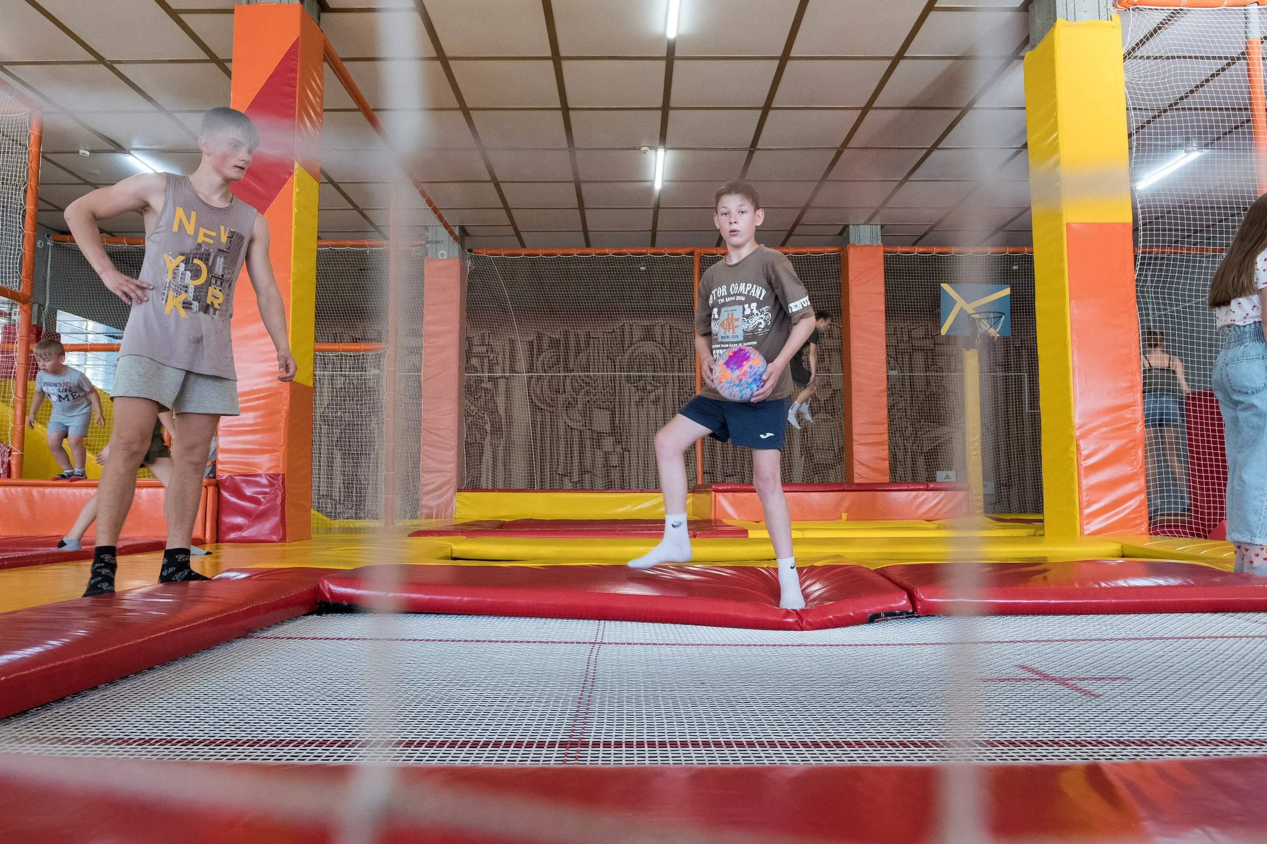 Enfants jouant dans un espace de trampoline intérieur avec des filets de sécurité, un garçon au centre tenant un ballon, d'autres enfants en arrière-plan.