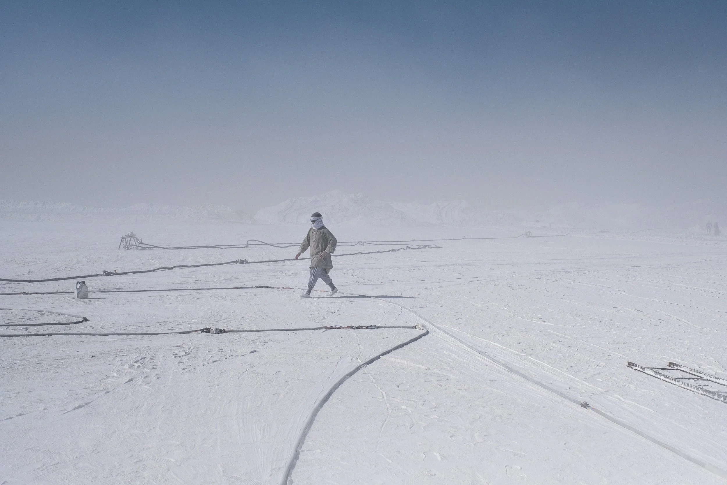 Personne marchant dans un paysage blanc avec des câbles électriques au sol, ciel dégagé et montagnes en arrière-plan.