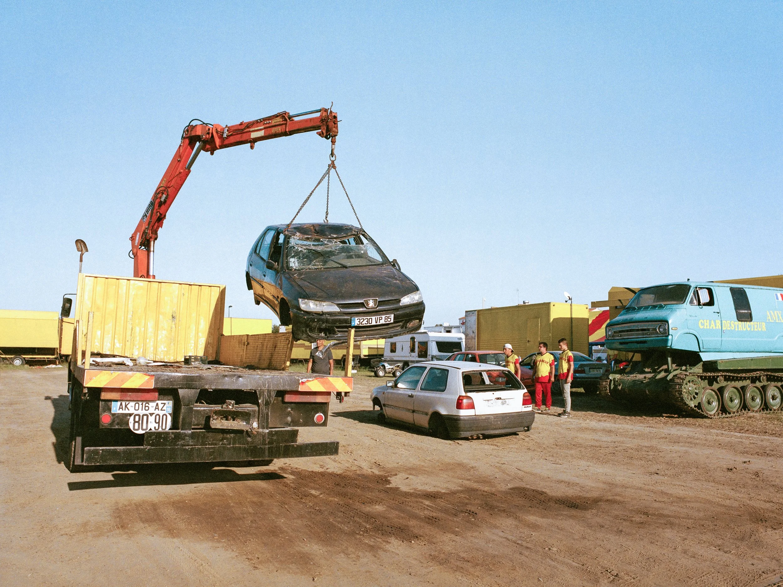 Une grue déplaçant une voiture endommagée lors d'un show de Monster Truck, avec des personnes en gilets de sécurité et un camion de remorquage.