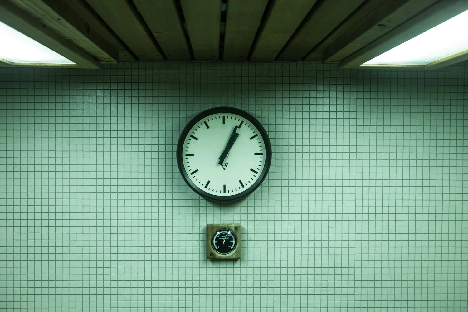 Horloge dans un sauna indiquant 12h02, avec un mur en carreaux blancs et un petit chronomètre en dessous