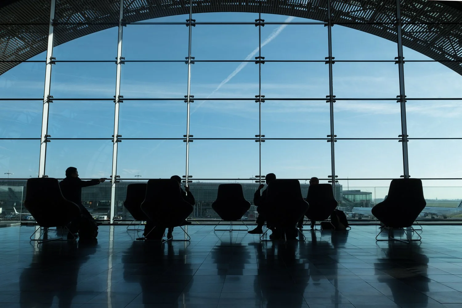 Vue intérieure de l'aéroport Charles de Gaulle avec des passagers en silhouette assis devant une grande fenêtre qui donne sur l'aéroport et le tarmac, avec des avions visibles à l'extérieur. 