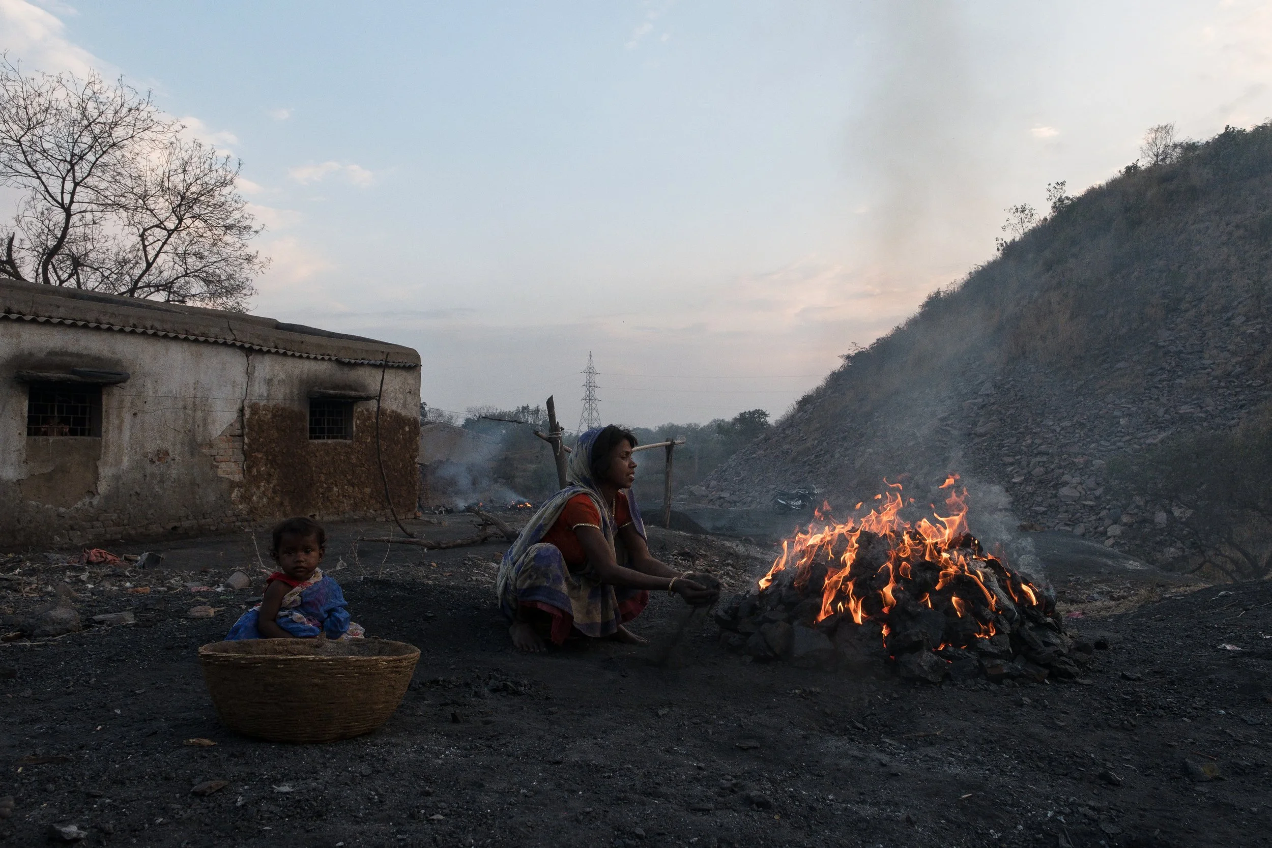 Une femme et un jeune enfant assis près d'un feu de charbon dans un environnement rural, avec une maison en ruine en arrière-plan et une colline rocheuse dans le fond, sous un ciel partiellement nuageux.