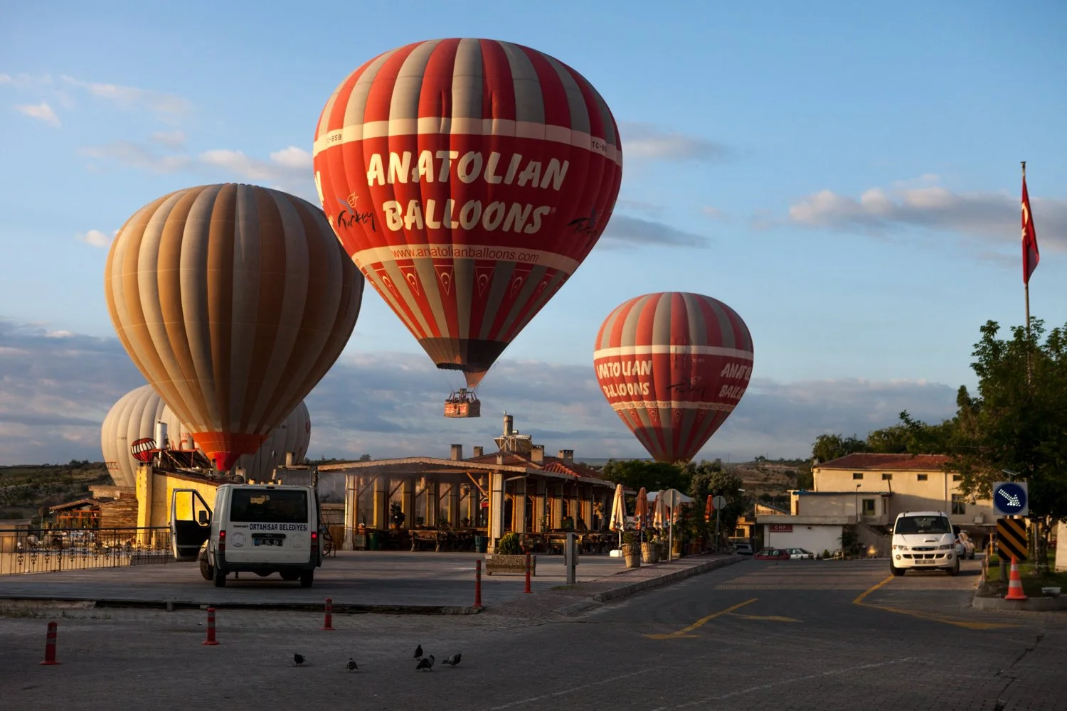 Plusieurs ballons dirigeables colorés flottent dans le ciel au-dessus d'un village avec des bâtiments, des voitures et un drapeau turc, en Cappadoce, Turquie