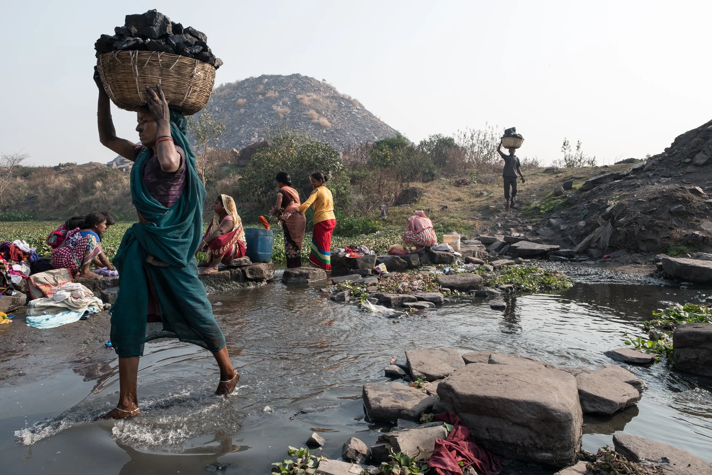Groupe de femmes et d'enfants lavant des vêtements dans une rivière, dans un paysage rocheux et montagneux, avec un ciel clair.