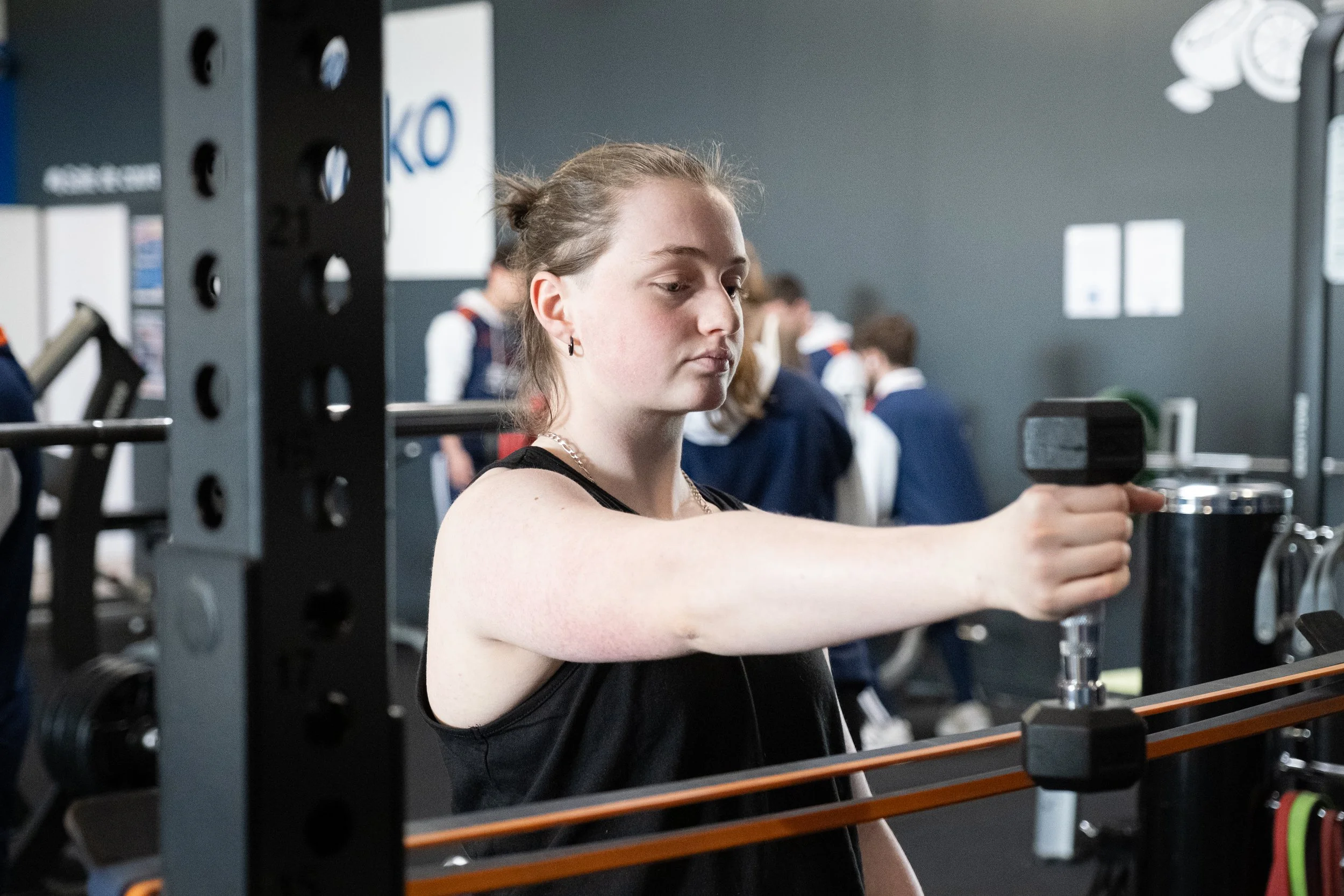 Jeune femme soulevant un haltère dans une salle de sport, avec d'autres personnes en arrière-plan.