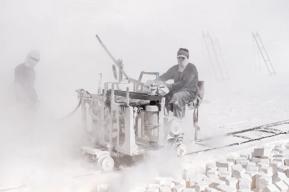 Deux personnes travaillant dans un environnement poussiéreux, utilisant un engin pour couper des blocs de pierre.