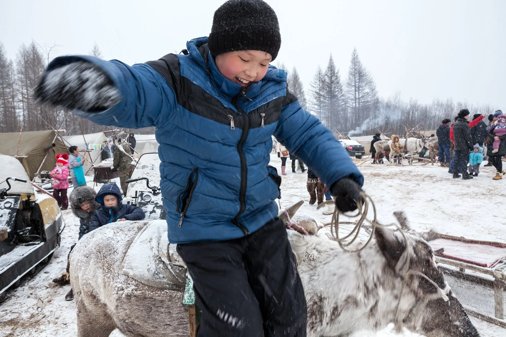 Un enfant en manteau bleu et bonnet noir saute du dos d'un renne lors d'une fête dans un environnement enneigé en Russie.