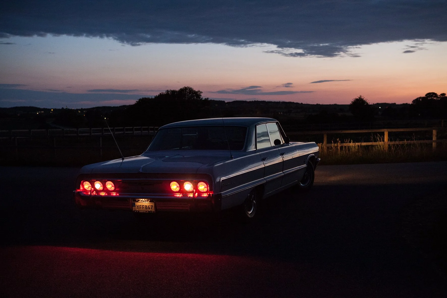 Voiture classique noire garée au coucher du soleil, avec un ciel crépusculaire et un paysage rural en arrière-plan.