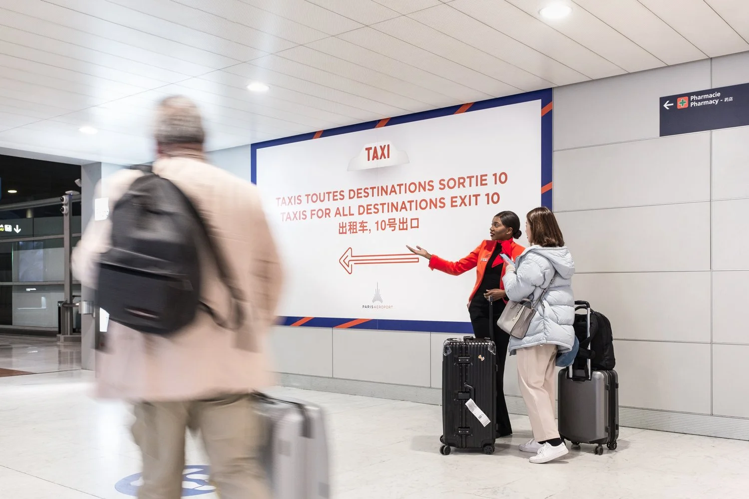 Un groupe de voyageurs attendant à un panneau d'information dans un aéroport. Le panneau indique que toutes les destinations de taxis sortent à la sortie 10, avec des instructions en français, anglais, chinois et japonais.